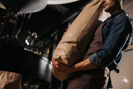 partial view of worker in apron holding paper bag of coffee beans in hands