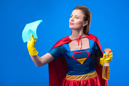 portrait of woman in superhero costume and rubber gloves with rag and detergent isolated on blueの写真素材