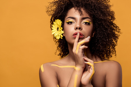 Young sensual african american woman with artistic make-up and gerbera in hair holding finger on...