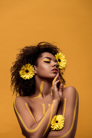 Young tender african american woman with artistic make-up and gerbera in hair isolated on orange backgroundの写真素材