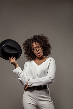 Young angry african american woman in white shirt wearing glasses holds hat isolated on grey backgroundの写真素材