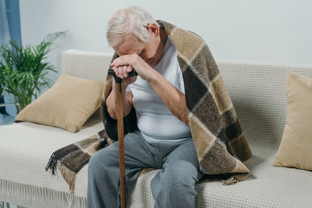 Tired senior man wearing plaid leans on a cane while sitting on sofaの写真素材