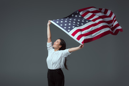happy young woman holding american flag in raised hands and looking away isolated on greyの写真素材