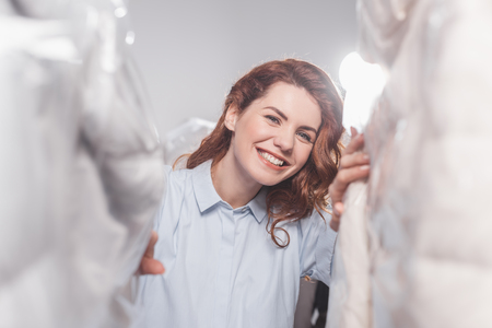 smiling female dry cleaning worker  looking at camera between clothing in plastic bags hanging at warehouseの写真素材