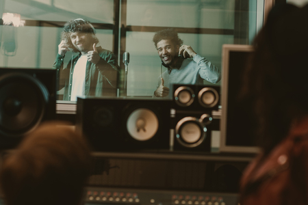 young handsome musicians recording song behind glass at studio and showing thumbs upの写真素材