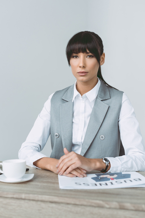 beautiful businesswoman sitting at table and looking at camera isolated on grayの写真素材