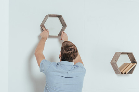 back view of young man hanging wooden bookshelf at homeの写真素材