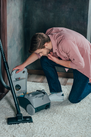 young man looking at broken vacuum cleaner at homeの写真素材