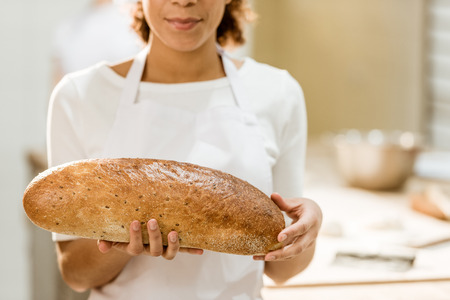 cropped shot of female baker holding fresh loaf of bread on baking manufactureの写真素材