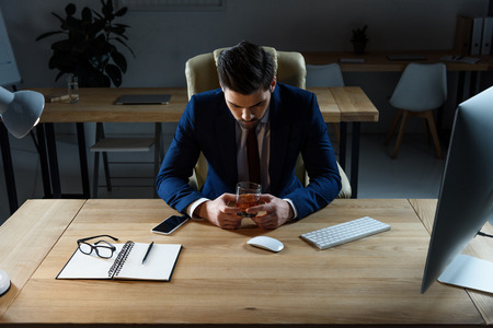 tired businessman looking at glass of whiskey in officeの写真素材