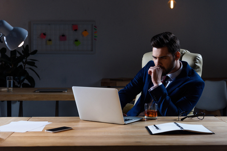 pensive businessman sitting in dark office and looking at laptopの写真素材