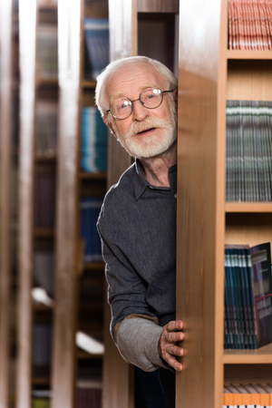happy grey hair librarian looking out from shelfの写真素材