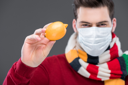 ill man in warm scarf and medical mask holding lemon, isolated on greyの写真素材