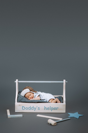 little baby in bodysuit lying in wooden toolkit with daddys helper lettering and hammer near byの写真素材