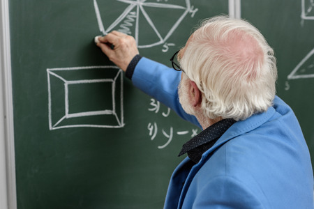 grey hair professor drawing with piece of chalk at blackboardの写真素材