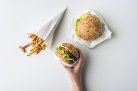 cropped view of female hand with hamburgers and french fries in paper cone, isolated on whiteの写真素材