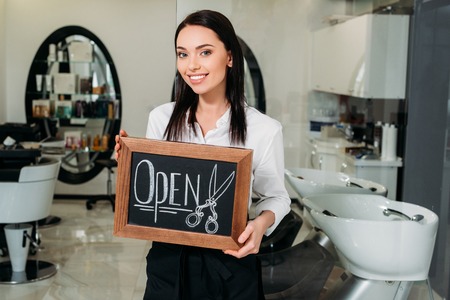 smiling brunette owner of salon standing with sign openの写真素材