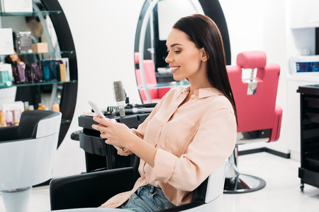 smiling woman sitting at hair salon and looking at smartphoneの写真素材