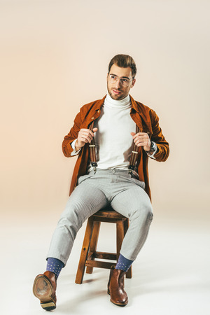 handsome pensive man looking away while sitting on wooden chair, on beigeの写真素材