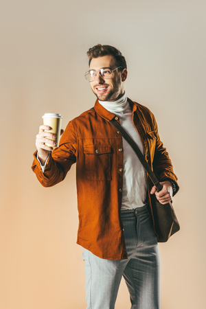 portrait of smiling man looking away and showing disposable cup of coffee in hand isolated on beigeの写真素材
