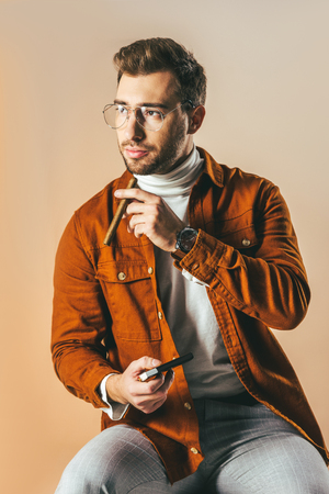 pensive stylish man with cigar in hand looking away isolated on beigeの写真素材