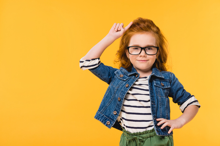 portrait of cute little kid in eyeglasses standing akimbo isolated on yellowの写真素材
