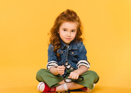 smiling child with eyeglasses in hands looking at camera isolated on yellowの写真素材