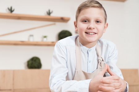 smiling boy in apron looking at camera while leaning on table at cafeの写真素材