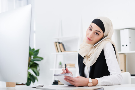 portrait of arabic businesswoman in hijab with notebook talking on smartphone at workplace in officeの写真素材