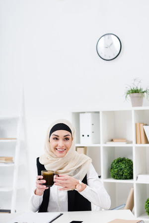 portrait of cheerful muslim businesswoman with cup of coffee at workplace in officeの写真素材