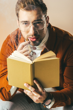 portrait of young man with cigar and book in hands looking at camera isolated on beigeの写真素材