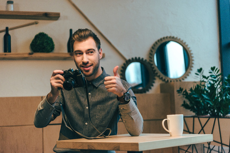 portrait of young man with photo camera showing thumb up in cafeの写真素材