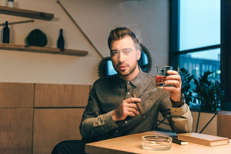 portrait of young man with glass of cognac and cigar in hands sitting at table in cafe