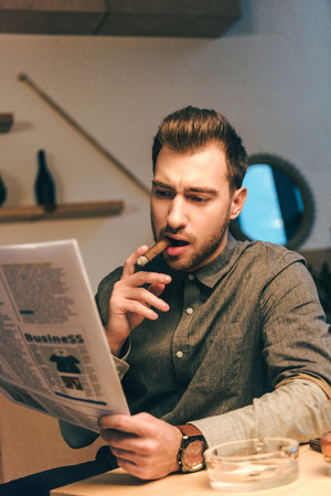 portrait of confident businessman smoking cigar while reading newspaper in cafeの写真素材