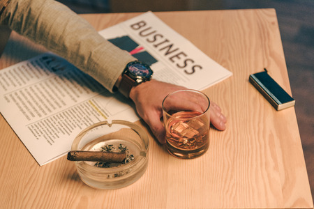 cropped shot of businessman sitting at table with glass of cognac, newspaper and cigar in ashtray in...