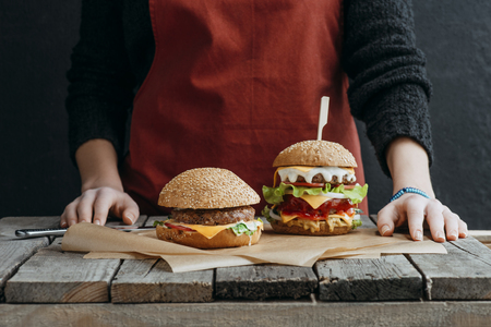 cropped view of girl in apron standing at wooden table with tasty cheeseburgers on baking paperの写真素材