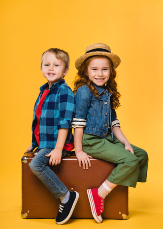 cheerful children sitting on leather suitcase isolated on yellowの写真素材