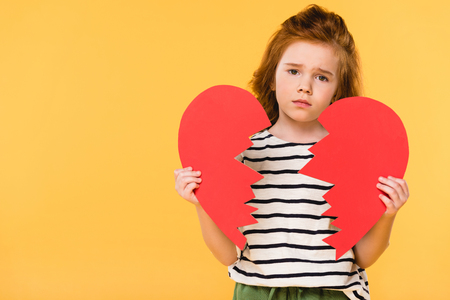 portrait of sad child with broken red paper heart isolated on yellow, st valentines day conceptの写真素材