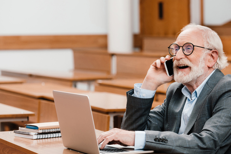 smiling grey hair professor sitting in empty lecture room and talking by smartphoneの写真素材