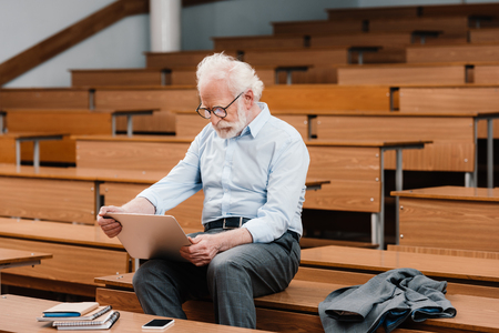 grey hair professor sitting on desk in empty lecture room and using laptopの写真素材