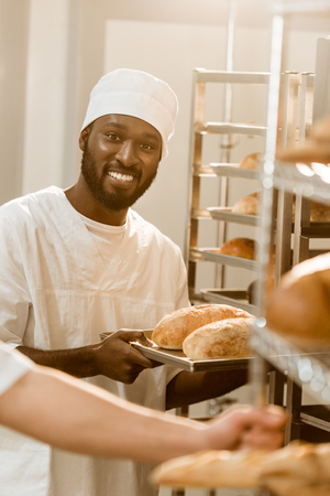 smiling african american baker holding loaves of bread on tray at baking manufactureの写真素材