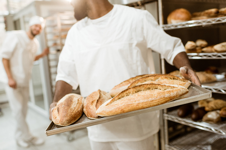 cropped shot of african american baker taking bread loaves from oven at baking manufacture