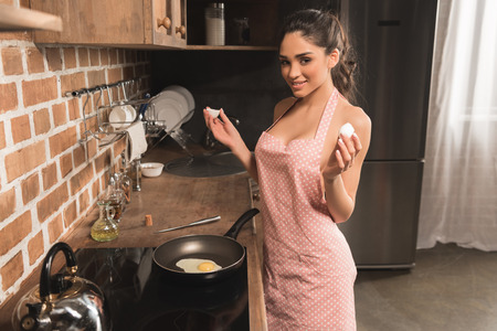sexy young woman in apron holding eggshell and smiling at camera while frying egg at kitchenの写真素材