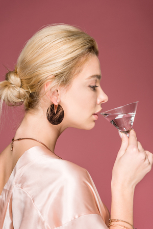 side view of attractive elegant girl in silk robe drinking cocktail, isolated on pinkの写真素材