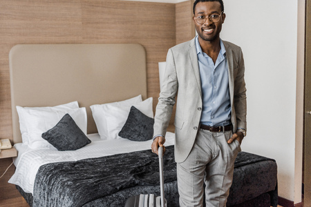 handsome smiling african american businessman with suitcase in hotel roomの写真素材