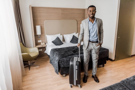 elegant african american businessman with suitcase in hotel room with bedの写真素材