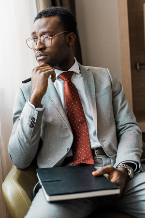 thoughtful african american businessman in gray suit sitting in armchair with journal in hotel roomの写真素材