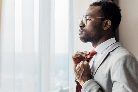 profile portrait of african american businessman adjusting red tie and looking at windowの写真素材