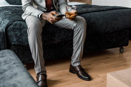 cropped view of african american businessman with glass of whiskey using smartphone in hotel roomの写真素材
