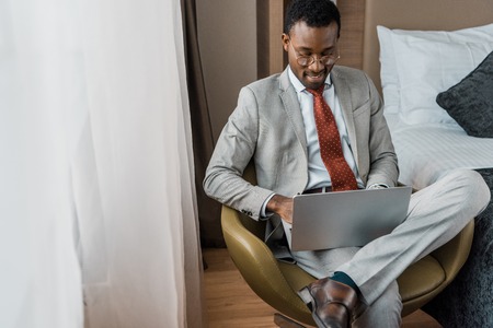 handsome smiling african american businessman working on laptop in hotel roomの写真素材
